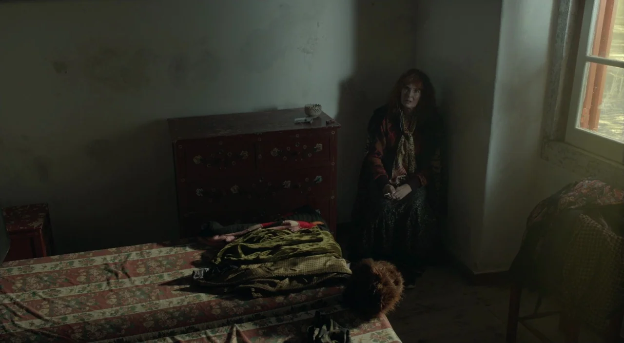 Cosmos (2015) movie still: medium — A sparsely furnished room with a painted wooden chest and a bed piled with fabric. A middle-aged woman sits in…