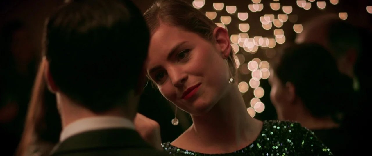The Canal (2014) movie still: medium — A dark, crowded event space features bokeh lights in the background. A woman in a green sequined dress looks…