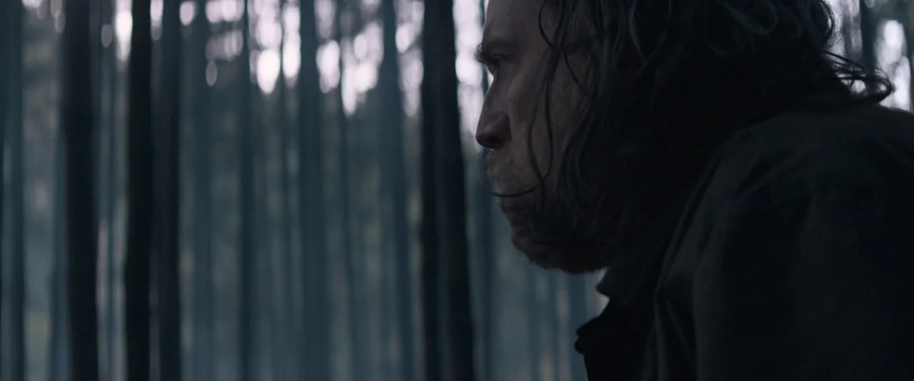 Pig (2021) movie still: closeup — A dense stand of slender pine trees forms a vertical, blurred background pattern. A man with unkempt dark hair and…