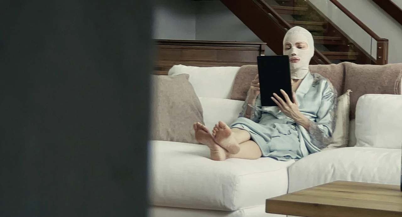 Goodnight Mommy (2022) movie still: medium — A minimalist, modern living room features a white sofa and a wooden staircase. A bandaged woman relaxes…