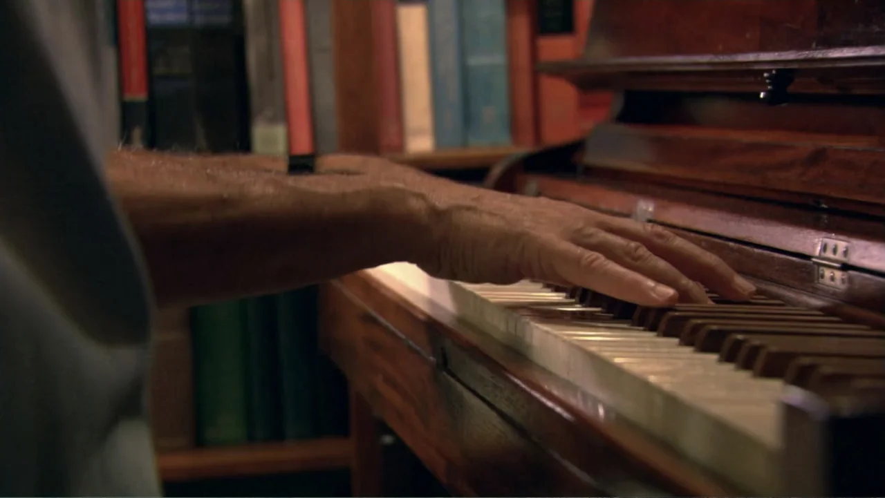 David McCullough: Painting with Words (2008) movie still: closeup — A close up view shows a wooden upright piano and a bookshelf filled with books.…