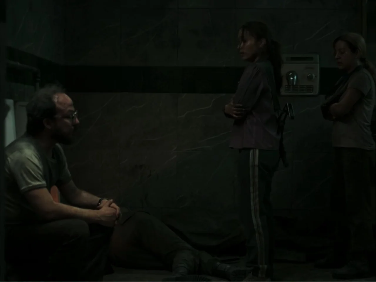 November (2025) movie still: medium — A dark, cramped room features dark-tiled walls and a small wall-mounted electrical unit. Three people are…