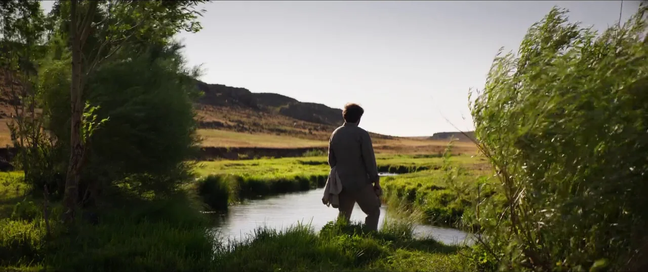 About Dry Grasses (2023) movie still: fullbody — A stream winds through a green grassy plain under a clear bright sky. A man stands on the bank…