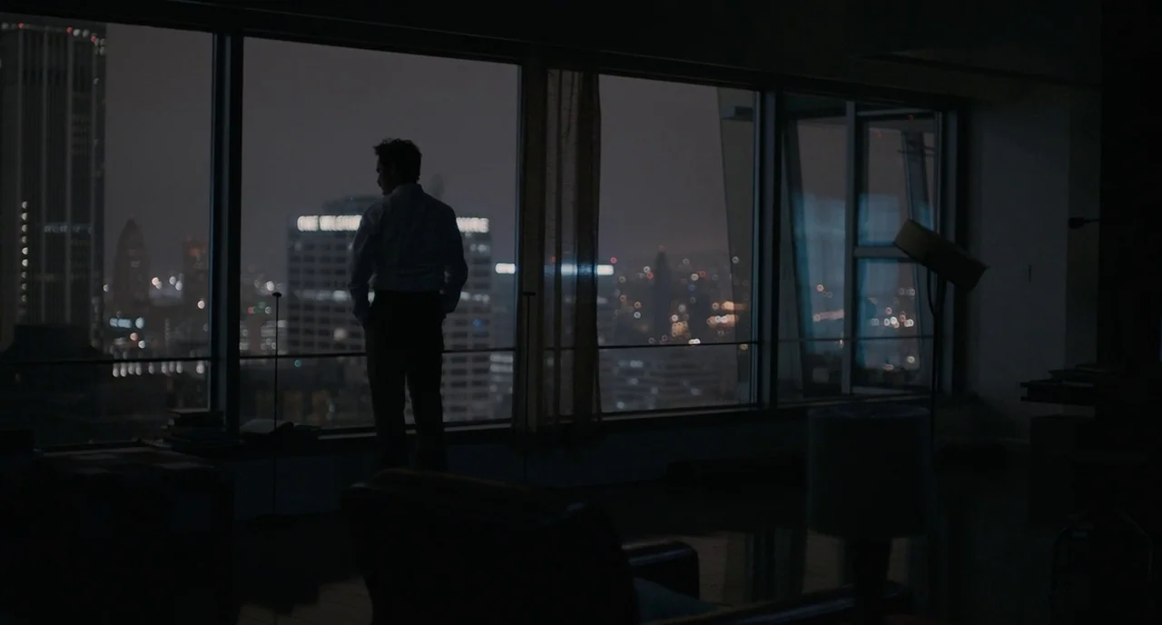 Her (2013) movie still: wide — A dark apartment interior at night features floor-to-ceiling windows overlooking a city skyline. A man stands facing…