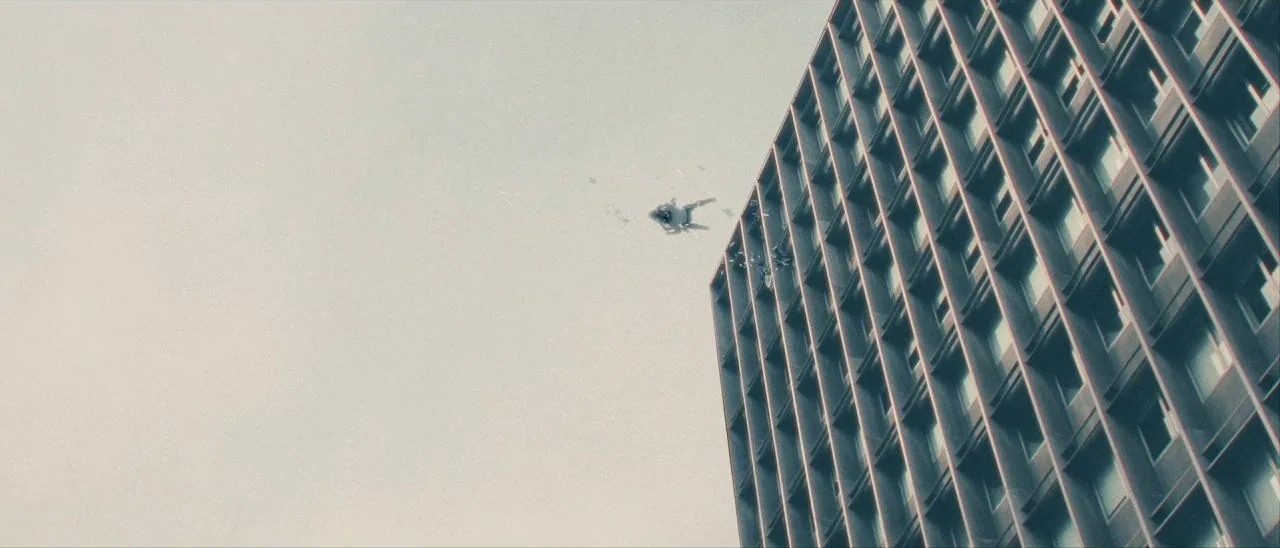 Archenemy (2020) movie still: wide — A tall modern office building with a repeating window grid stands against a grey sky. A small figure and debris…