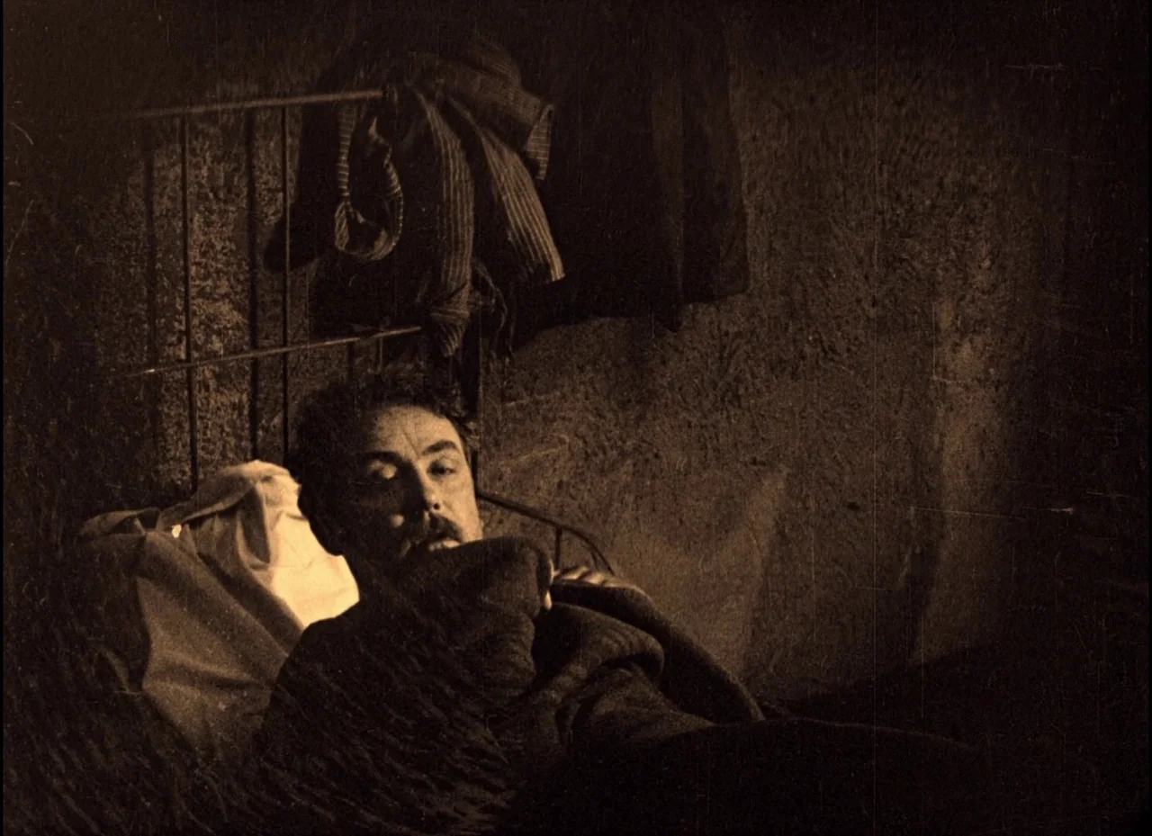 The Phantom Carriage (1921) movie still: medium — A stone-walled interior space features a metal bed frame against the wall. A man lies in the bed…