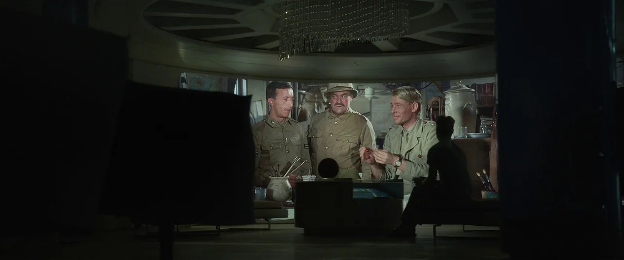 Prometheus (2012) movie still: medium — A dimly lit room features a chandelier and mid-century furniture. Three men in military uniforms converse…