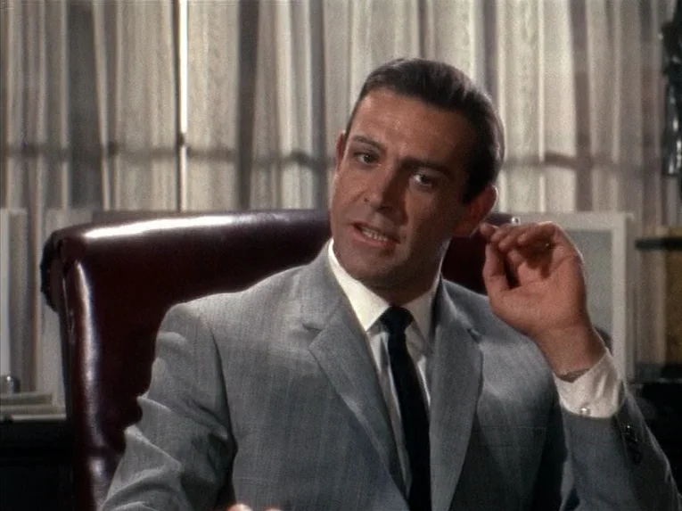 Marnie (1964) movie still: medium — A man sits in a professional setting with vertical window blinds in the background. He is wearing a grey suit…