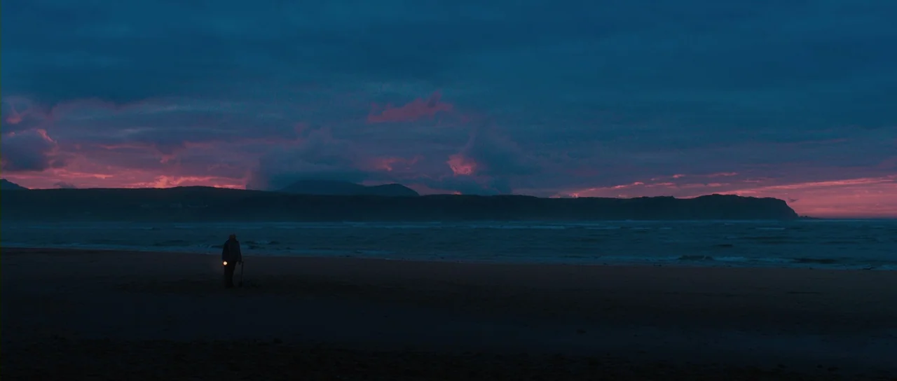 Grabbers (2012) movie still: wide — A dark sand beach stretches out toward a distant coastline under a twilight sky. A lone man stands at the…