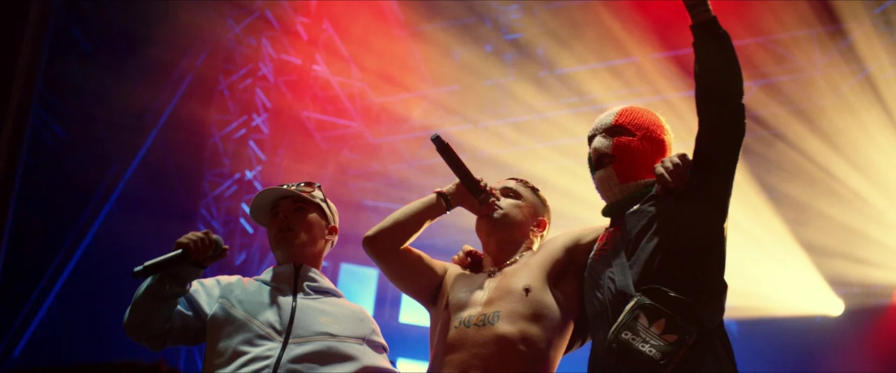 Kneecap (2024) movie still: medium — Three men stand together on a brightly lit concert stage with exposed truss architecture. The central figure…
