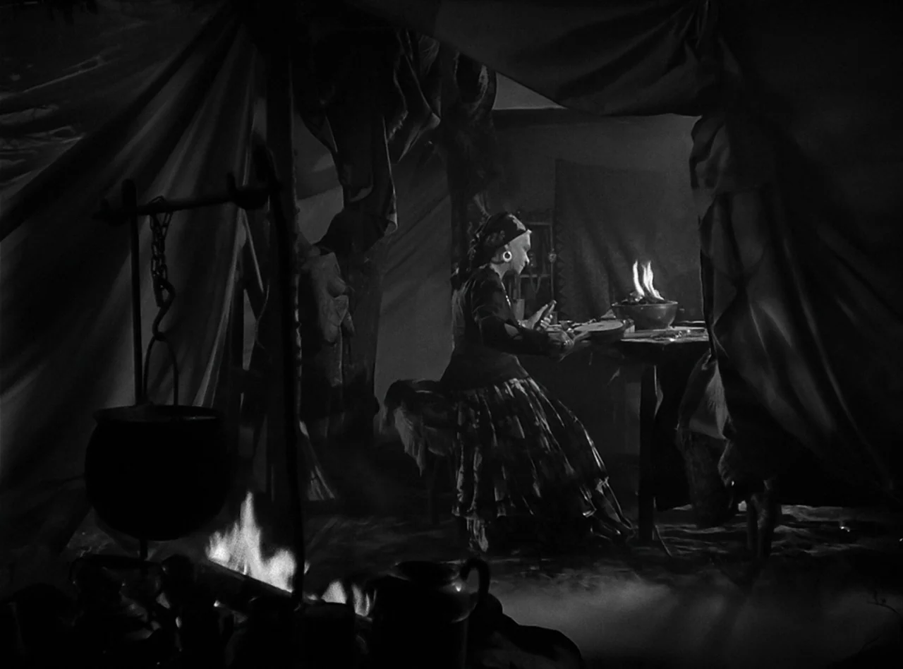 The Wolf Man (1941) movie still: wide — A dimly lit interior of a fabric tent with low-hanging ceiling drapes. A woman sits at a wooden table beside…