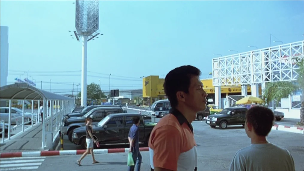Tropical Malady (2004) movie still: medium — A sunny, expansive parking lot area with various parked vehicles and a large billboard. Two men stand…