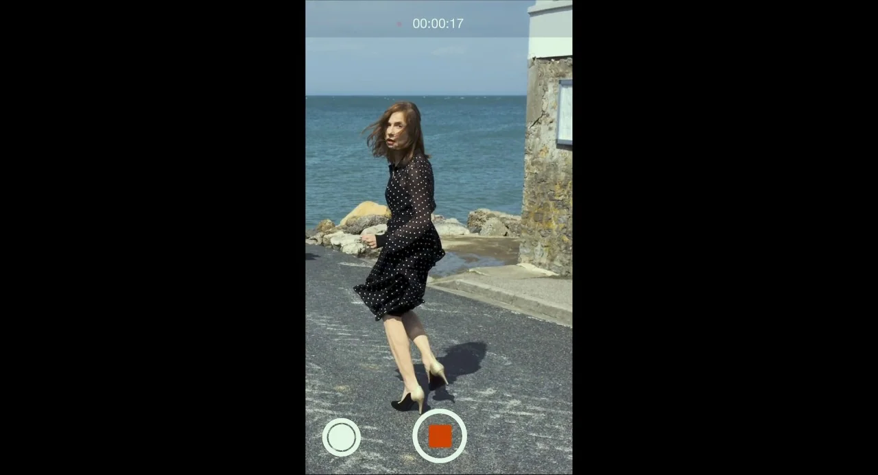 Happy End (2017) movie still: fullbody — A paved coastal path runs alongside the sea with stone embankment walls. The woman walks on the asphalt…