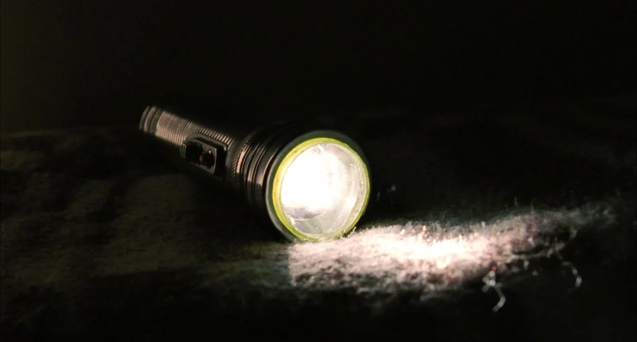 Incendies movie still: closeup — A metallic flashlight lies horizontally on a dark, rough-textured surface. The device projects a focused beam of…