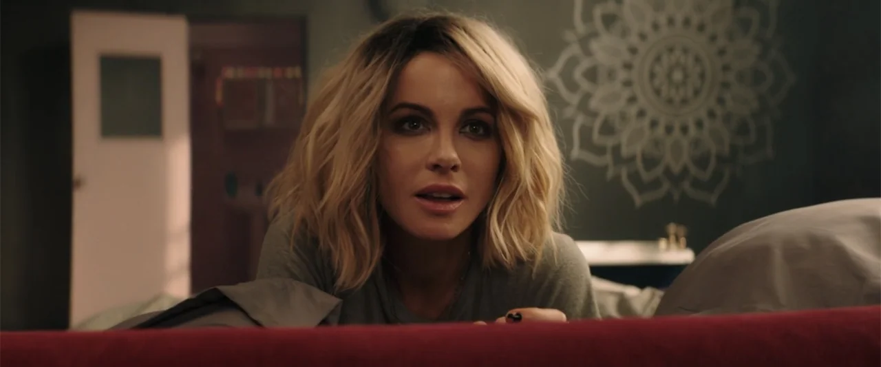 Jolt (2021) movie still: closeup — A modern bedroom with a dark wall featuring a decorative white circular mandala design. A woman lies prone on a…