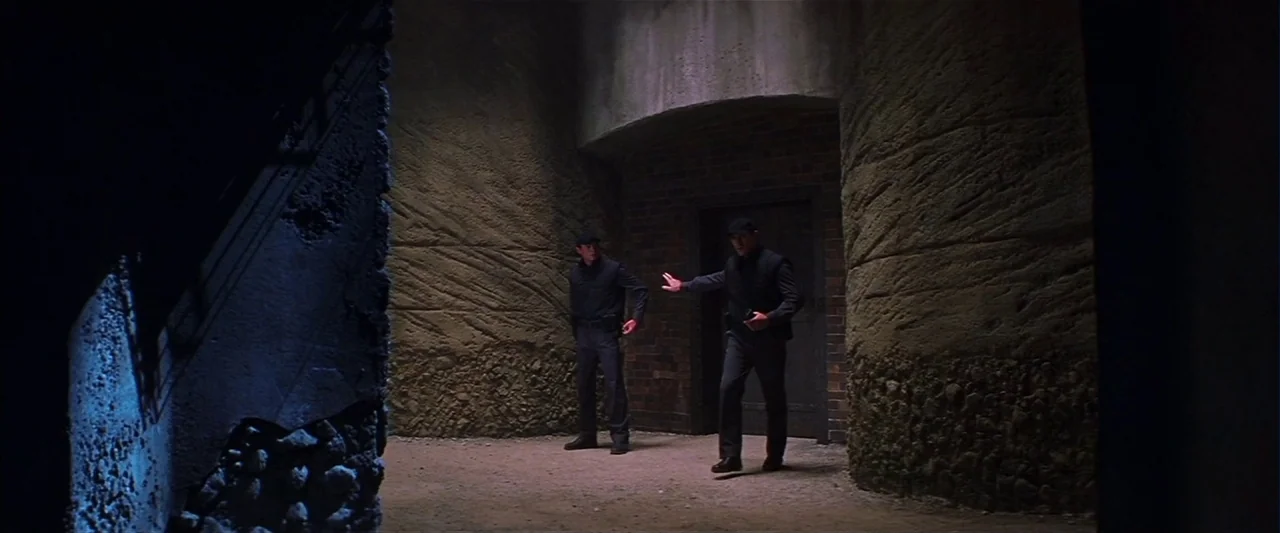 Mission: Impossible II (2000) movie still: wide — A dark subterranean chamber features curved stone walls and a recessed central brick doorway. Two…