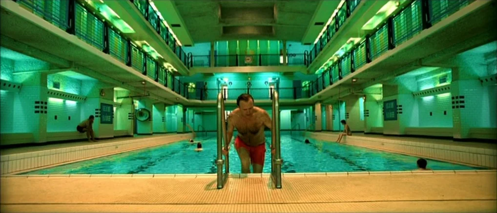 Amelie (2001) movie still: fullbody — An indoor public pool facility with balconies, tiled walls, and rows of lockers. A man in red swim trunks…