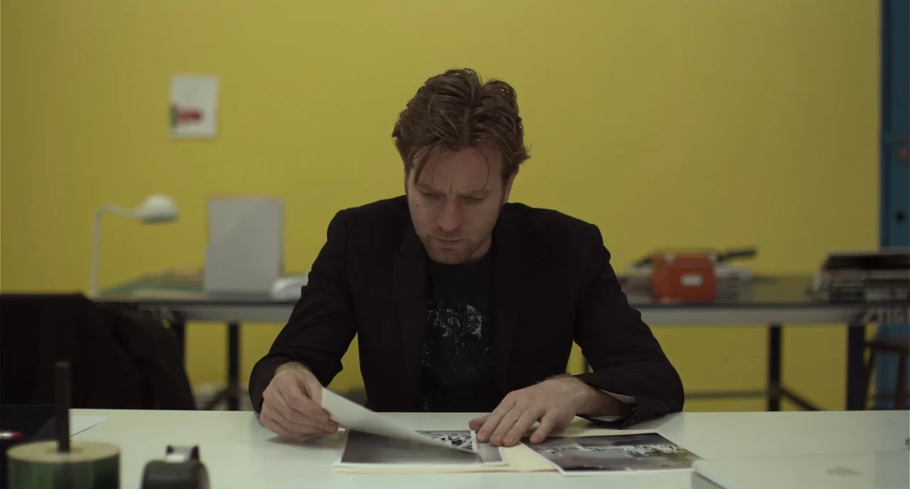 Beginners (2010) movie still: medium — A man sits at a white desk against a vibrant yellow wall in a room. The man examines several black and white…