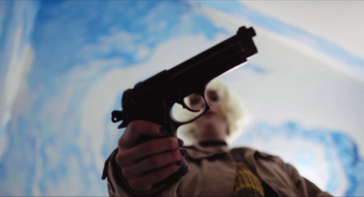 Like Me movie still: closeup — A person stands in a room with blue and white cloud-patterned wallpaper. The subject holds a black handgun extended…