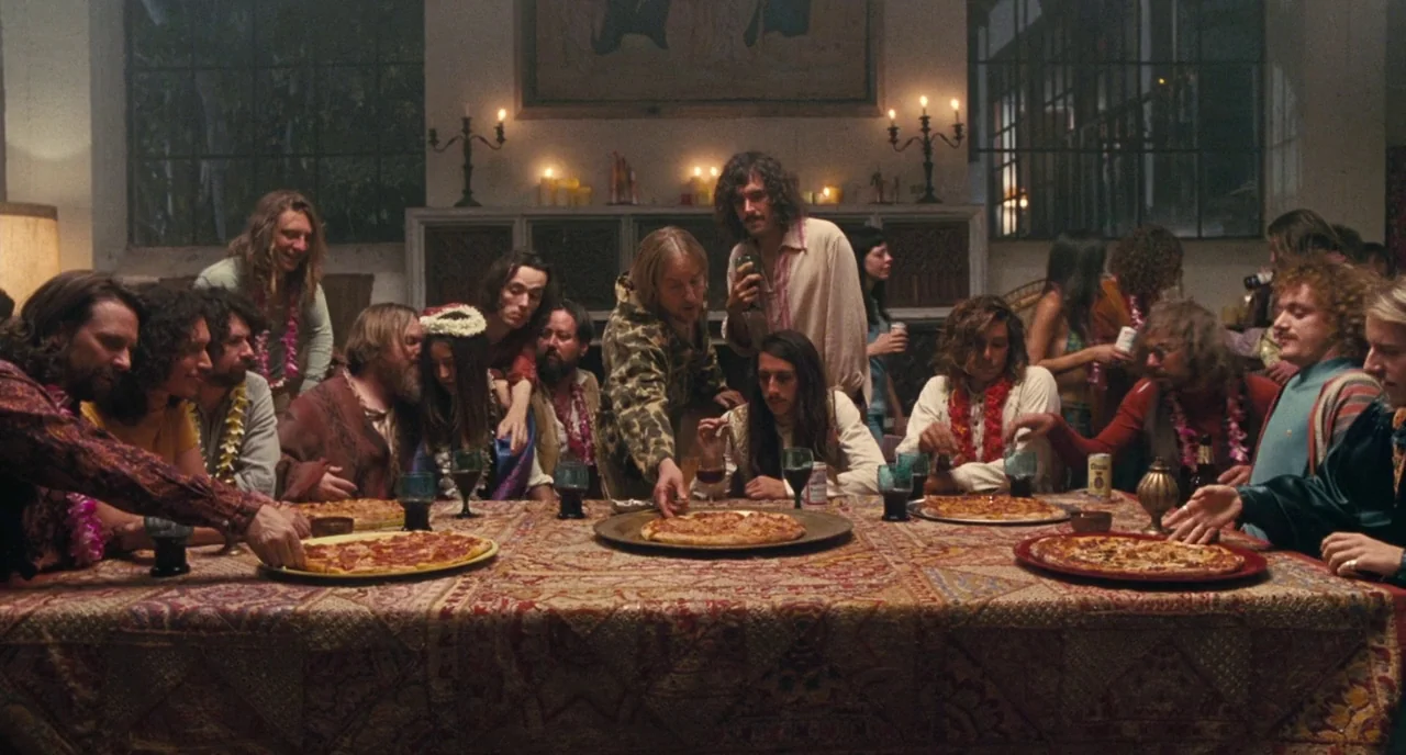 Inherent Vice (2014) movie still: wide — A group of people are seated around a large table covered in a patterned tablecloth. They are sharing…