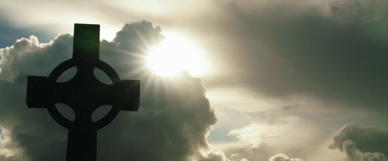 The Banshees of Inisherin (2022) movie still: wide — A stone Celtic cross stands against a bright, cloudy afternoon sky. Sun rays flare from behind…