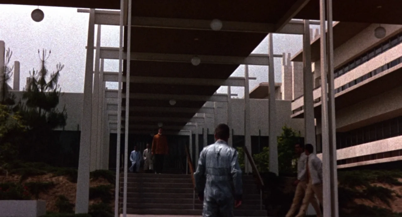Queen of Blood (1966) movie still: wide — A modernist brutalist complex features a long concrete walkway supported by white pillars. A man walks up…