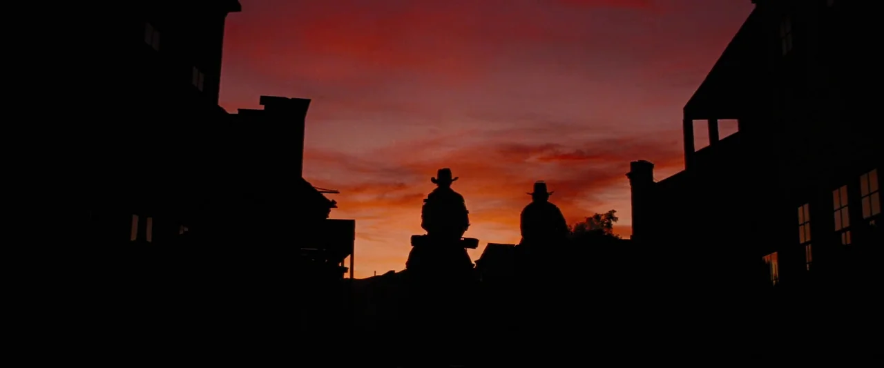 Django Unchained (2012) movie still: wide — A quiet western town street with wooden buildings framing a brilliant sunset sky. Two riders on…
