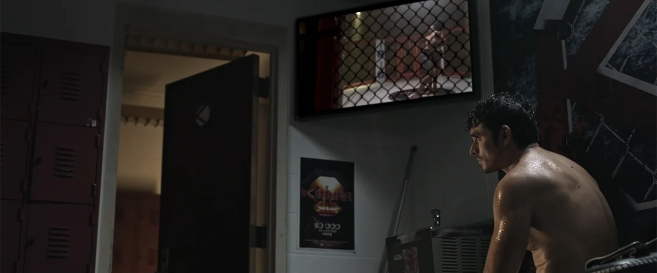 Kimura (2017) movie still: medium — A dimly lit locker room features red metal lockers and a wall-mounted television screen. A shirtless man sits on…