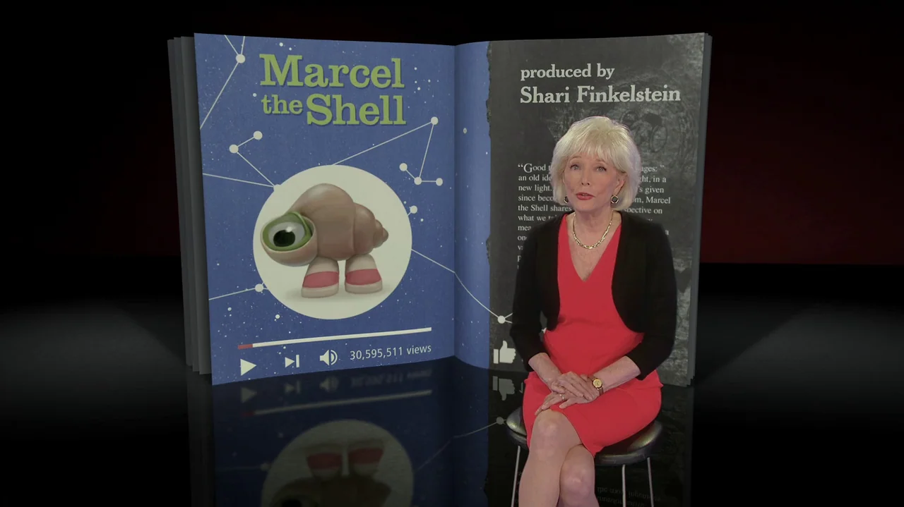 Marcel the Shell with Shoes On (2021) movie still: medium — An elderly woman sits in front of an open, digital book displaying video graphics. The…