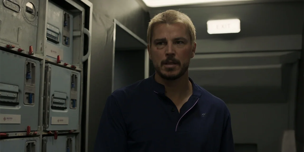 Fight or Flight (2025) movie still: medium — An airplane galley shows metal storage compartments and an illuminated overhead exit sign. A man with…