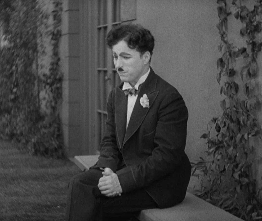 City Lights (1931) movie still: medium — A stone bench sits against a stucco wall covered with climbing vines. A man in a suit sits on the bench…