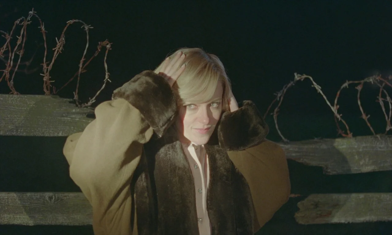 Spencer (2021) movie still: medium — A woman stands in front of a wooden fence lined with barbed wire at night. She holds both hands to her hair…