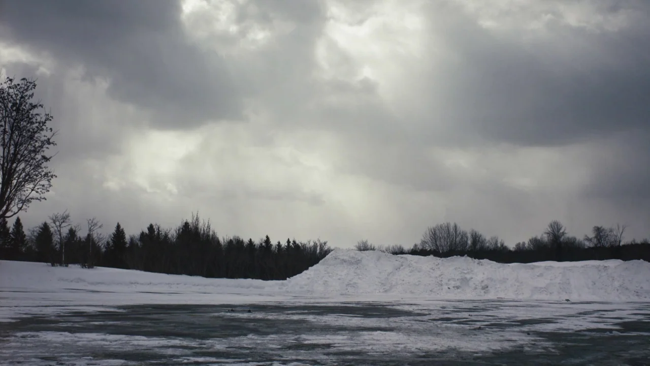 The Blackcoat’s Daughter (2015) movie still: establishing — A flat asphalt surface is partially covered in patches of dirty melting snow. Large…