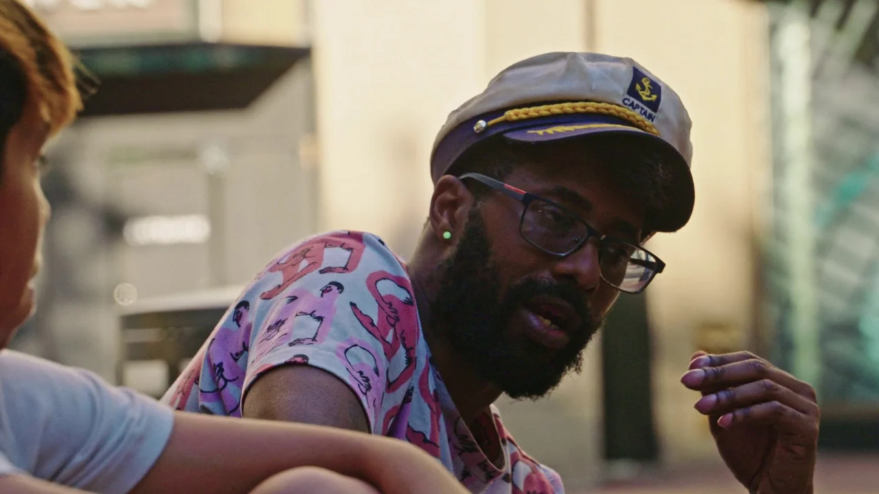 Gasoline Rainbow (2023) movie still: medium — An outdoor urban setting with a blurred building facade in the background. A man wearing a captain's…