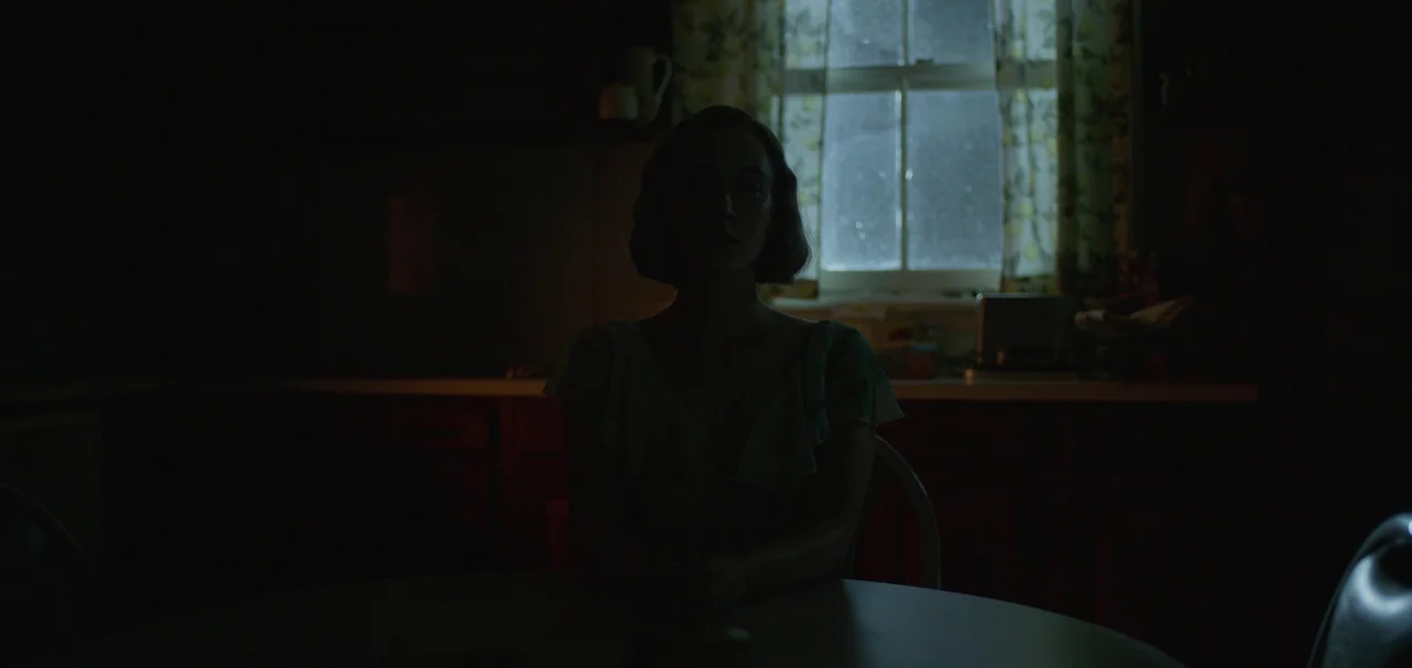 Foe (2023) movie still: medium — A dark, sparsely lit interior space with a window covered by patterned curtains. A woman sits in shadow at a table…