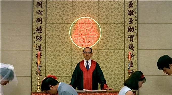 A Summer at Grandpa’s (1984) movie still: medium — A formal wedding ceremony takes place before a wall featuring traditional gold scroll banners. A…