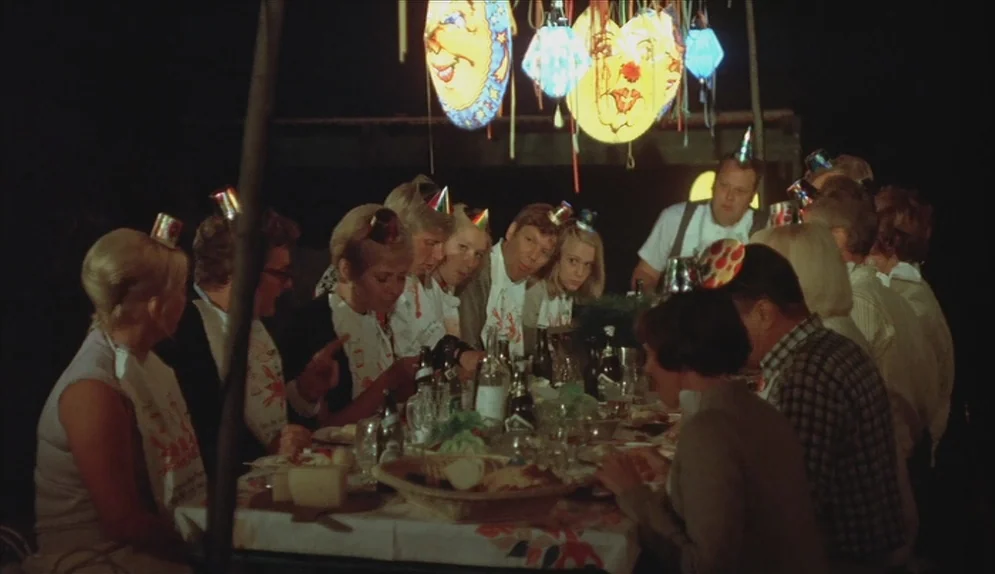 A Swedish Love Story (1970) movie still: medium — A group of adults sits at a long table outdoors at night under lanterns. They are wearing paper…