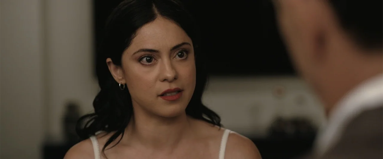 Play Dirty (2025) movie still: closeup — A dimly lit indoor space provides a backdrop to a conversational exchange. A woman with dark hair looks…