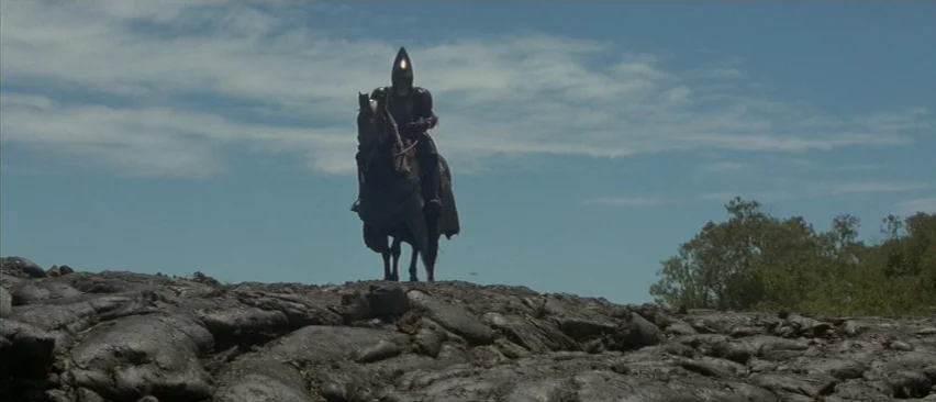 Planet of the Apes (2001) movie still: wide — A dark armored rider on horseback stands atop a rough volcanic rock formation. A clear blue sky with…