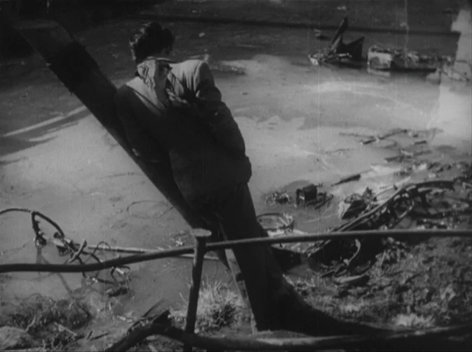 Drunken Angel (1948) movie still: fullbody — A desolate area covered in scattered metallic wreckage and debris. A man in a suit leans against a…