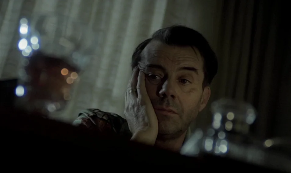 Despair (1978) movie still: closeup — A dimly lit room with heavy curtains in the background and a dark foreground. A middle-aged man rests his face…