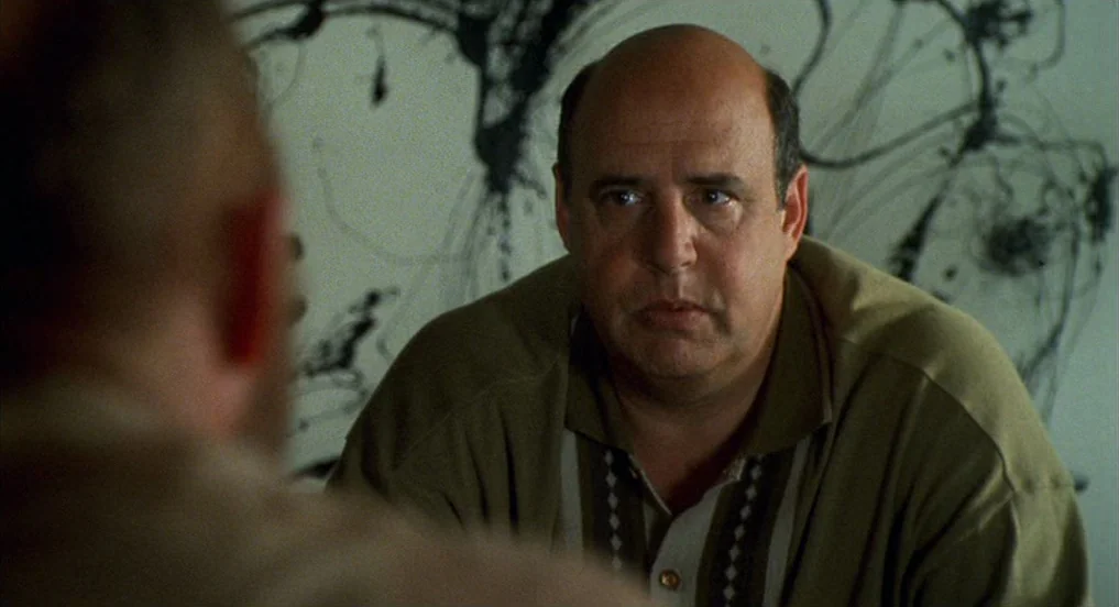 Pollock (2000) movie still: medium — A man sits before an abstract black-on-white painted mural on a white wall. The man wears a knit shirt and…