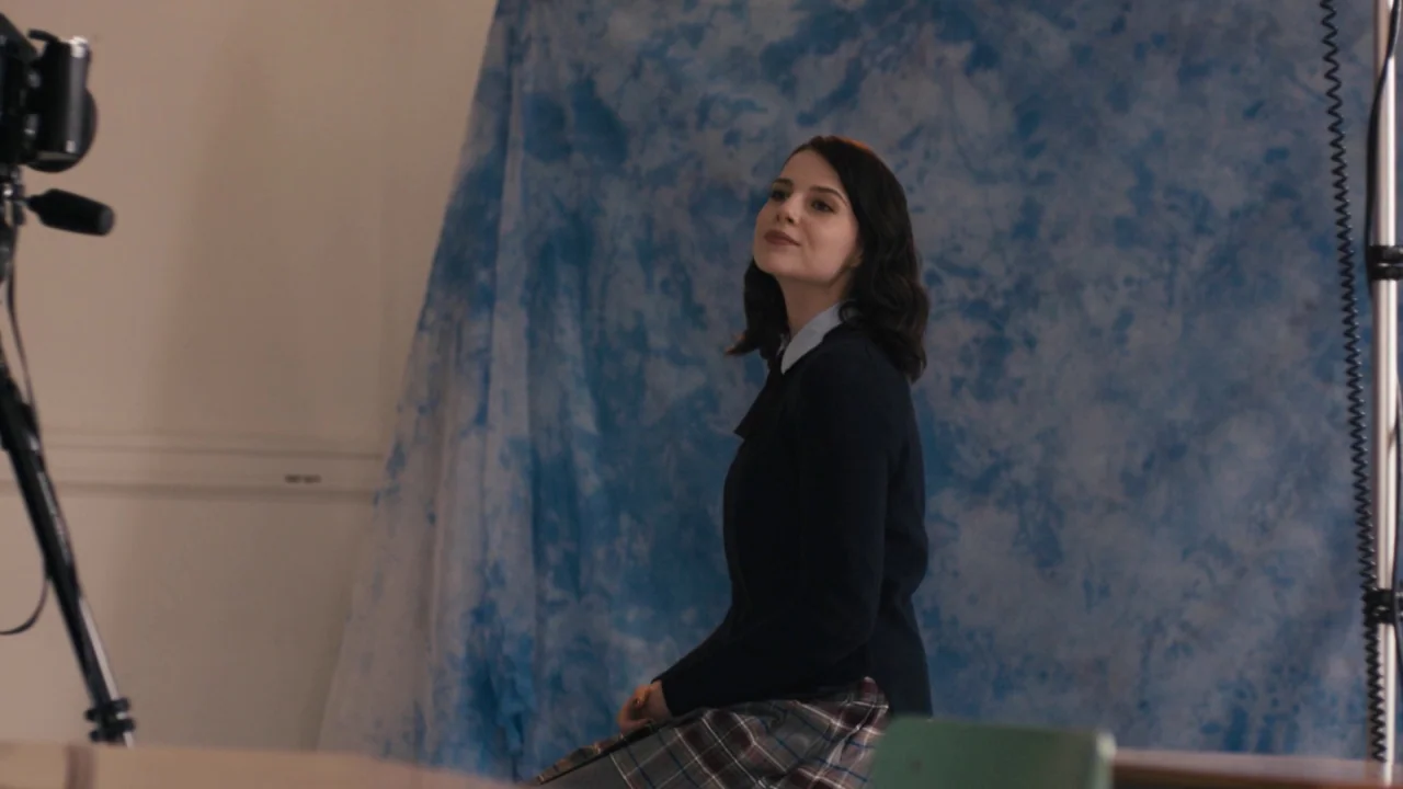 The Blackcoat’s Daughter (2015) movie still: medium — A student sits in front of a blue mottled photography backdrop in a room. She is positioned…