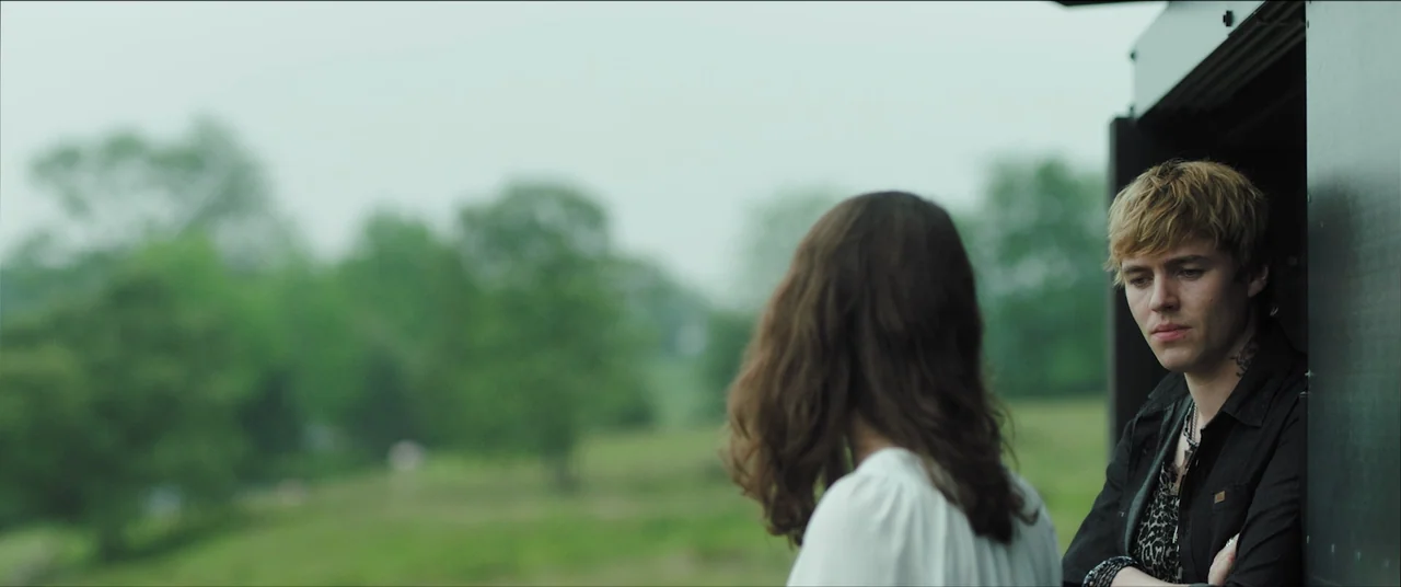 The Feast (2021) movie still: medium — An open grassy area with soft-focus trees stands behind two people in the frame. The woman is shown from…