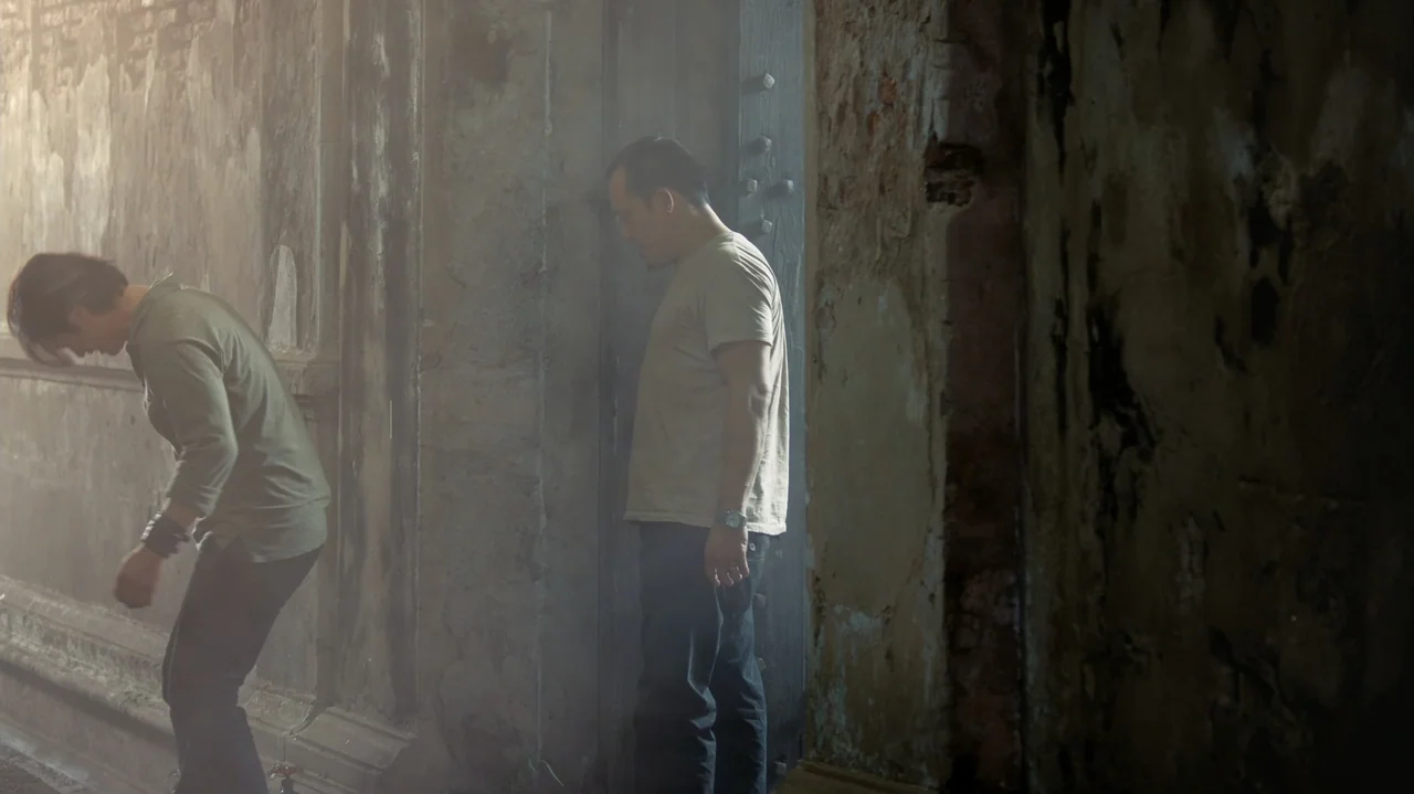 Zero Tolerance (2015) movie still: fullbody — A dusty narrow hallway features stone walls and a wooden door frame. One man leans into a room while…