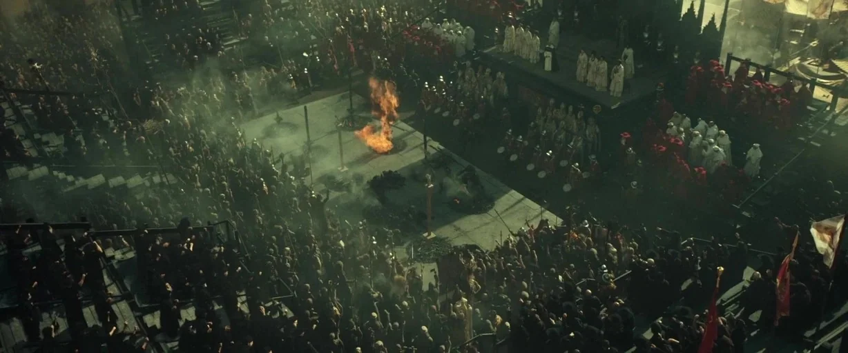 Assassin’s Creed (2016) movie still: establishing — A large stone plaza is filled with dense crowds surrounding a central fire pit. Spectators stand…