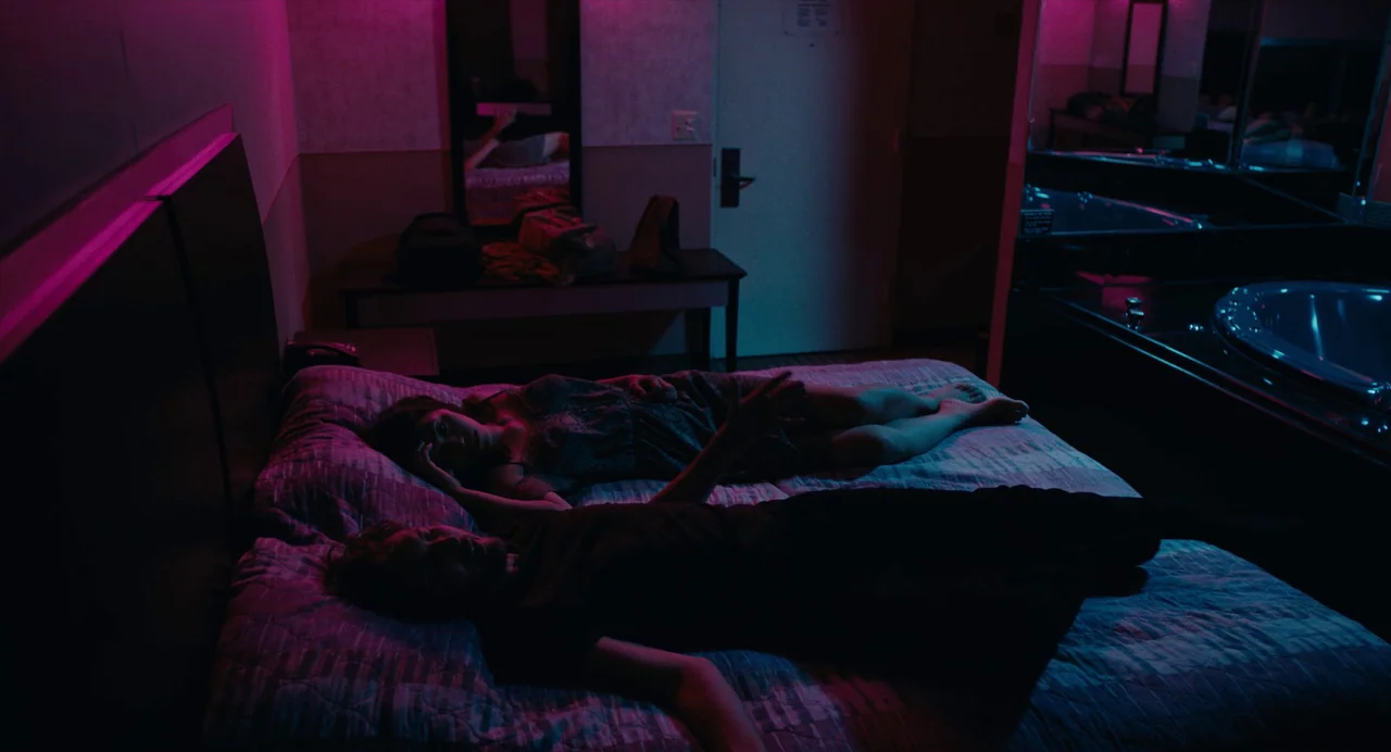 Lingua Franca (2019) movie still: wide — A dark hotel room is illuminated by vibrant neon pink and blue light strips. A young man and woman lie…
