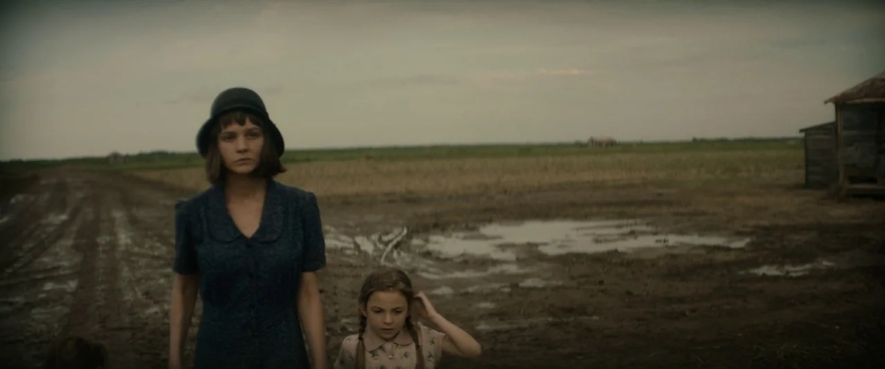 Mudbound (2017) movie still: medium — A flat, muddy rural landscape stretches out under a grey, overcast sky. An adult woman and a child walk toward…