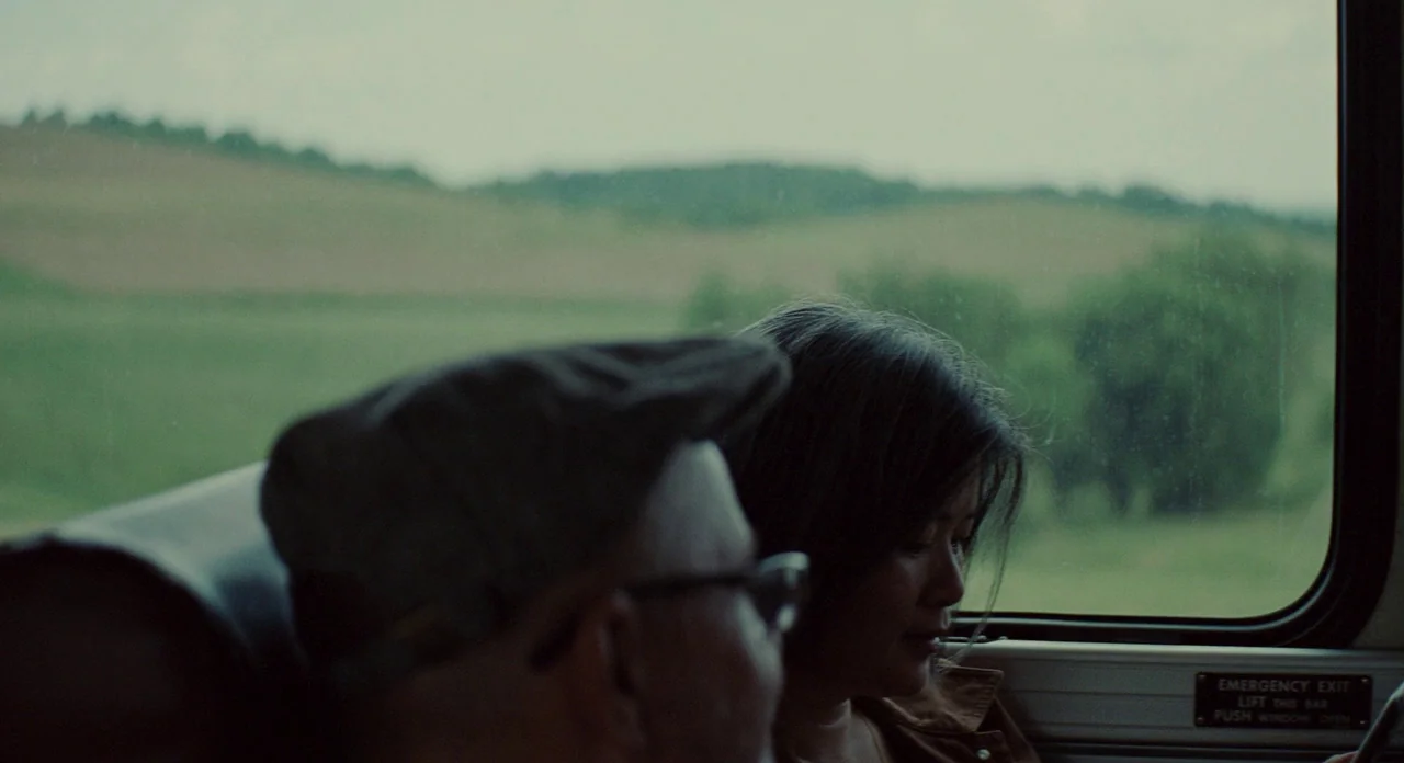 Promised Land (2012) movie still: medium — A view inside a passenger train with a window looking out at green hills. An older man wearing a cap and…