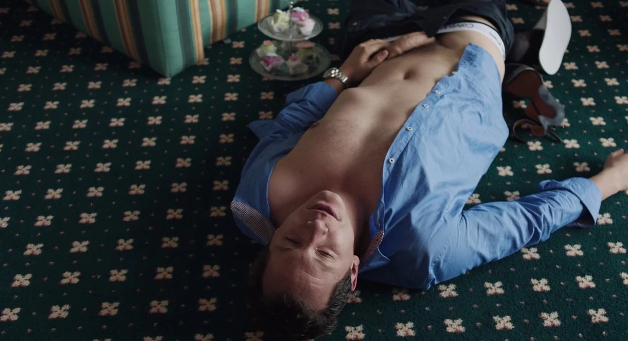 Toni Erdmann (2016) movie still: medium — A man lies on a dark green floral-patterned carpet inside a hotel room. Nearby sits a tiered stand…